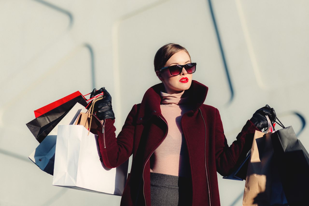 who-we-are Stylish young woman in sunglasses carrying shopping bags outdoors. Elegant fashion statement.